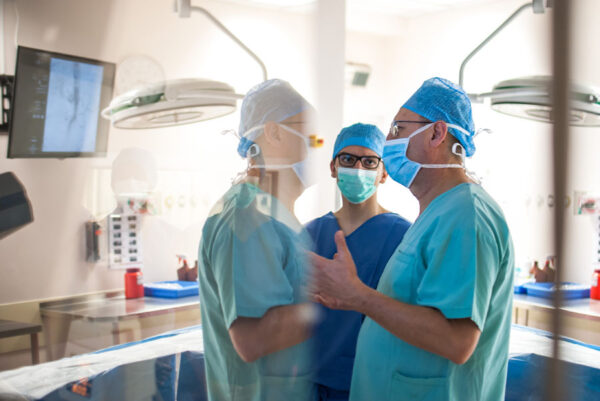 Surgeons in blue scrubs and masks discussing something in a modern operating room with reflections visible on a glass surface.