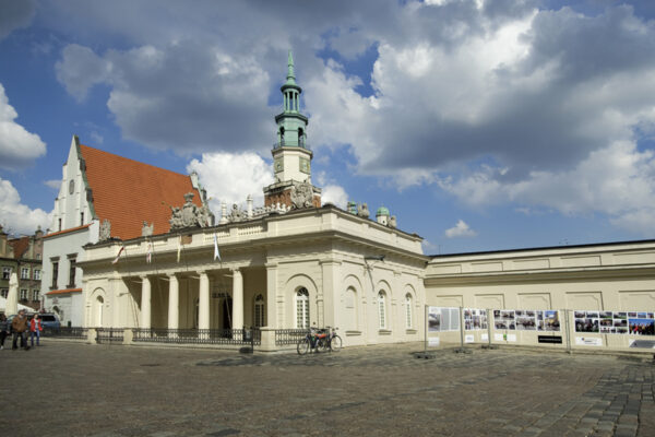 A historical building with a classic architectural design, featuring a tall tower with a greenish dome and a series of arches. The building is situated in a public square with cobblestones, and the sky is partly cloudy, giving the scene a timeless atmosphere.