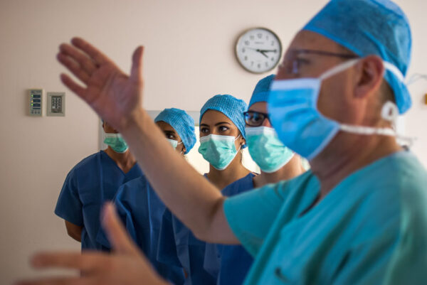 A group of healthcare workers in an operating room, dressed in surgical scrubs and masks. They appear to be receiving instructions or discussing a procedure, with one person gesturing while the others attentively listen, reflecting teamwork in a high-stakes environment.