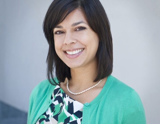 Sadhana Dharmapuri, M.D. A smiling professional woman wearing a green cardigan and a patterned dress. She has dark hair styled into a neat bob and stands against a plain background, exuding warmth and approachability.