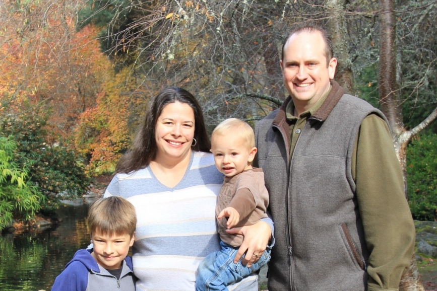 A family stands together smiling in front of a serene pond, capturing a joyful moment in nature.