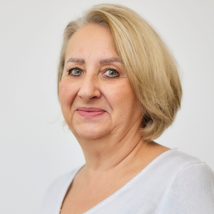 A portrait of a mature woman with blonde hair, wearing a white top. She is smiling softly with a gentle expression, set against a plain light-colored background.