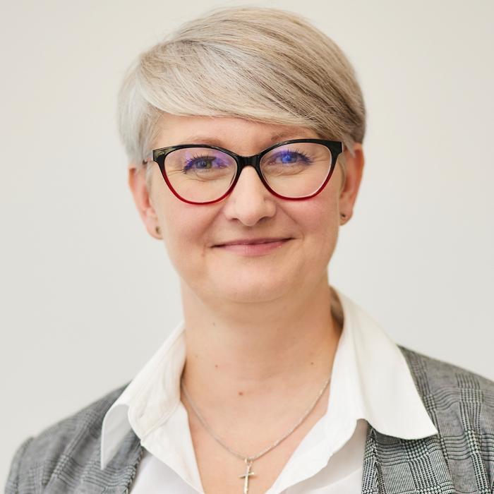 A professional-looking woman with short grey hair and glasses, wearing a light checkered jacket over a white shirt, smiling at the camera.