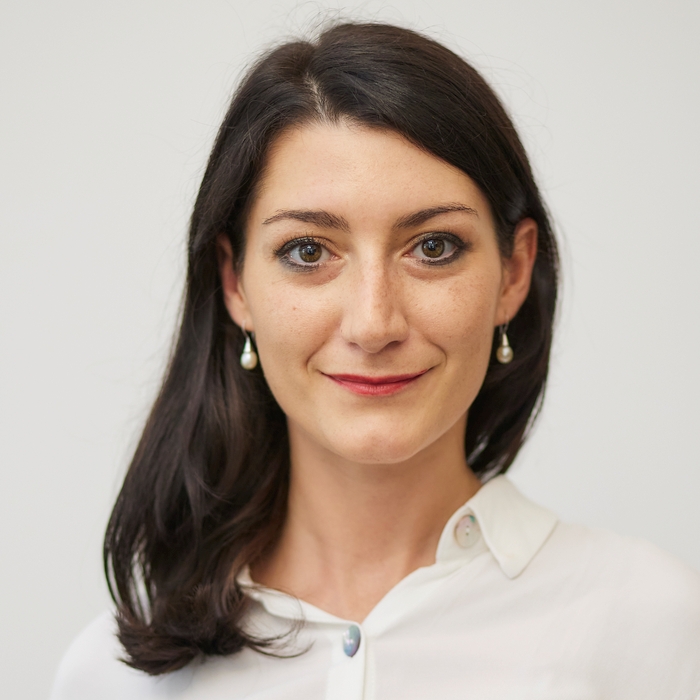 A woman with dark hair wearing a white shirt, standing confidently against a neutral background.