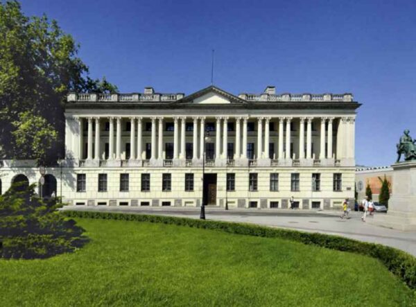 A grand neoclassical building with a row of tall columns along the facade, likely a government or significant public institution. The building is surrounded by a well-maintained lawn, and a few people are visible walking nearby, highlighting the stately and formal atmosphere of the location.