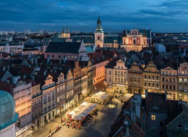 An evening aerial view of a vibrant European city square with historical buildings lit up, showcasing cafes and a bustling street atmosphere.