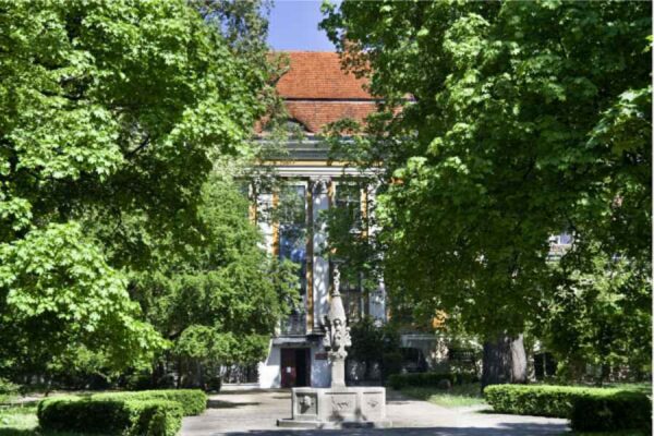 A museum entrance is partially obscured by large, leafy trees, with a statue visible in the foreground.
