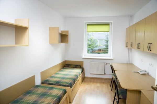 A small but cozy dormitory room with two beds, a desk, and storage cabinets, featuring a window with a green curtain and wooden flooring.