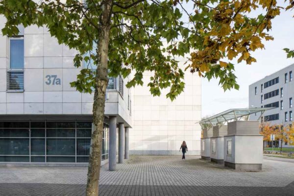 A modern university building with a clean, white facade marked as '37a.' A lone figure walks along a paved courtyard lined with autumn trees. The scene evokes a quiet academic atmosphere.