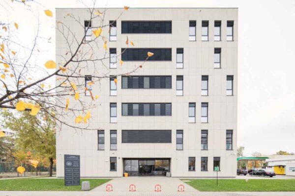 A symmetrical white educational building with tall windows and minimalistic design. The entrance is marked by a central open space, flanked by bike racks. The setting is modern and professional, with a hint of seasonal autumn colors from nearby trees.