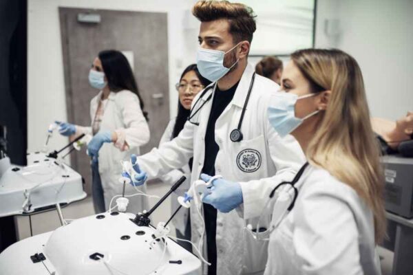 Medical students in lab coats, wearing masks and gloves, practicing laparoscopic surgery techniques on simulation machines. They are focused and working in a collaborative environment.