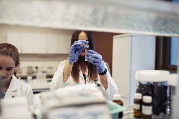 A student examines a vial of liquid in a lab while another student works beside her. The lab is filled with shelves and equipment for advanced scientific research.