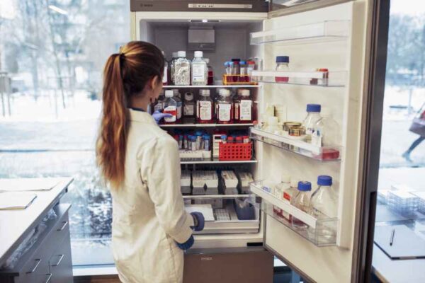 A student in a lab coat reviewing the contents of a well-stocked refrigerator filled with bottles, vials, and samples. The scene emphasizes careful inventory management in a research environment.