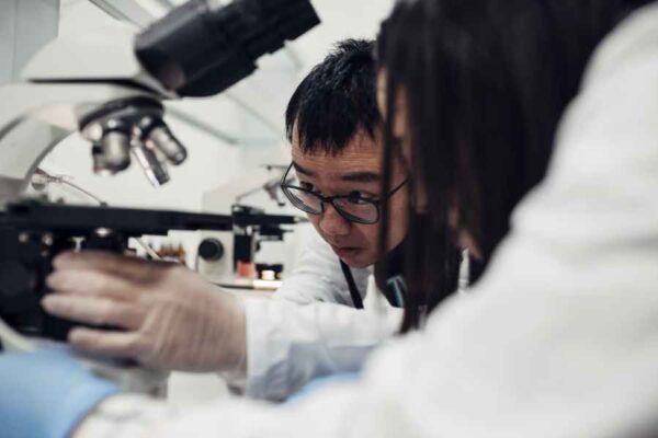 Two students, one male and one female, intensely focusing on looking through microscopes in a laboratory setting. The concentration on their faces suggests they are analyzing samples.