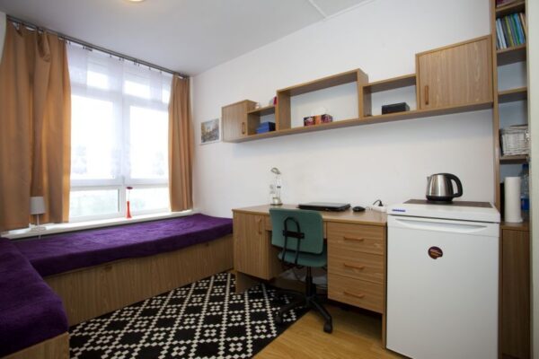 A cozy, single-person dorm room with a neatly arranged bed near a large window. The room features wooden furniture, including a desk, bookshelf, and a small refrigerator, creating a warm and functional living space.